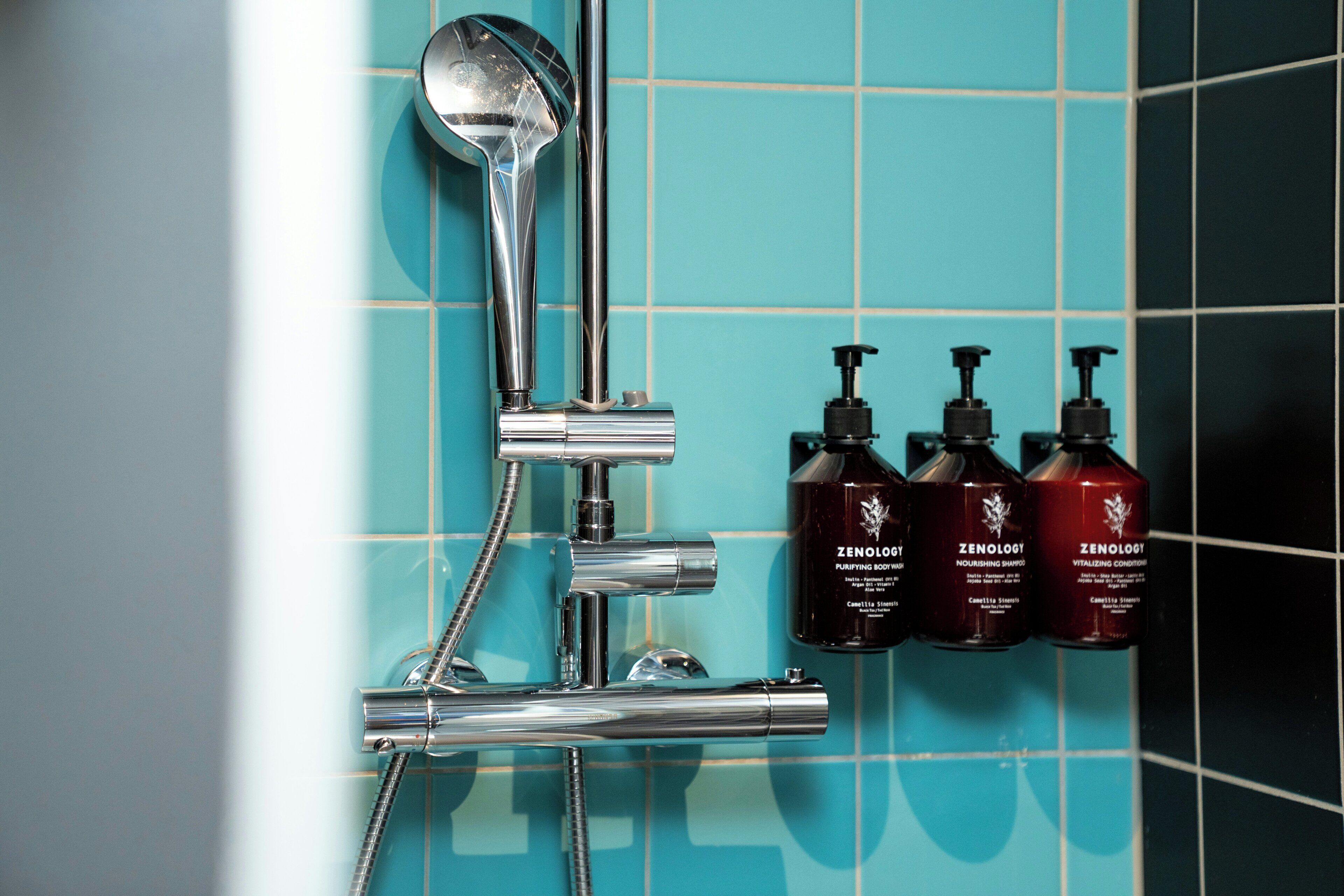 Un cabezal de ducha cromado, una ducha de mano y tres botellas de productos Zenology sobre una pared de azulejos.