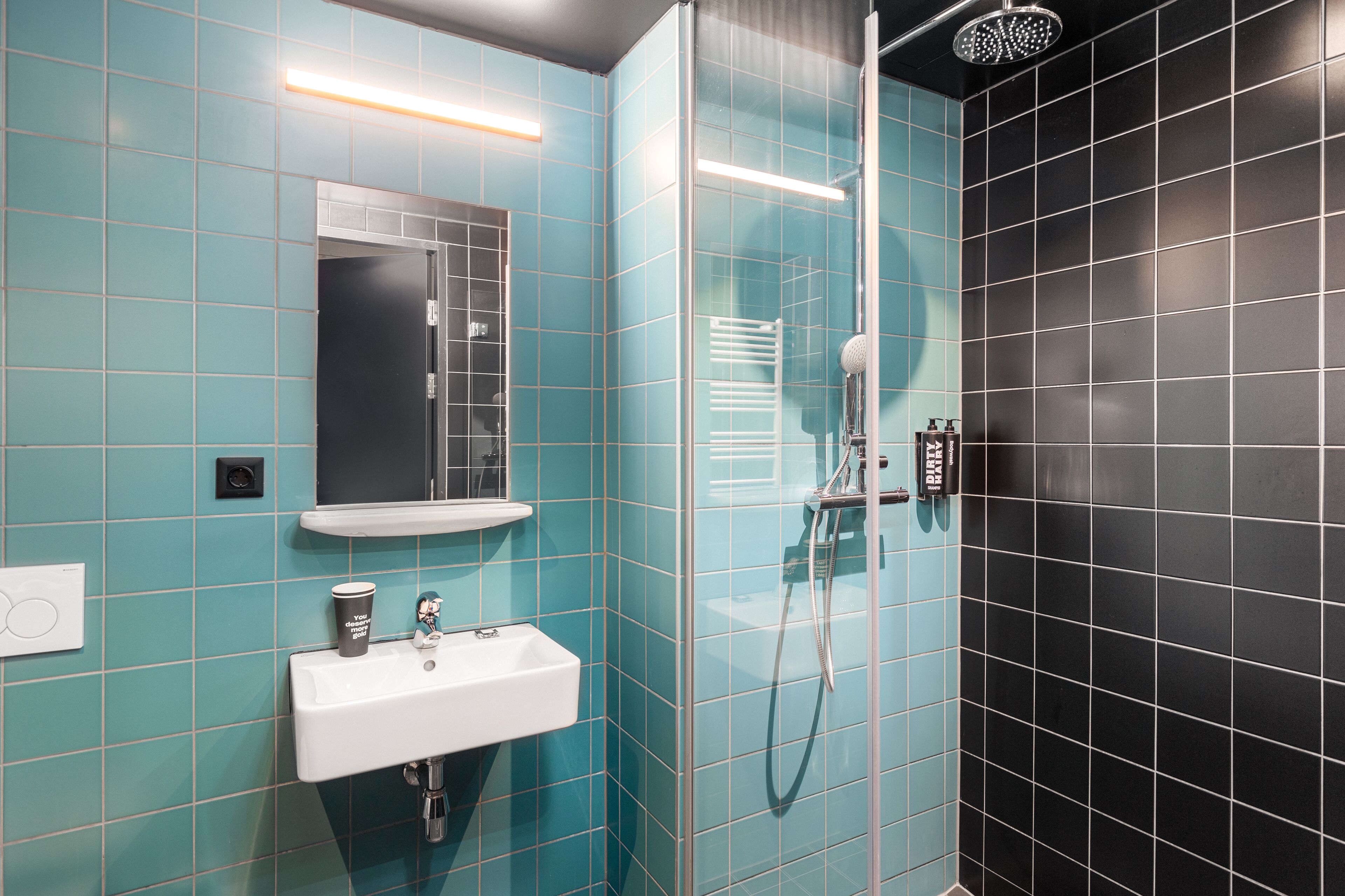 Un baño con un lavamanos blanco, un espejo, una ducha con cabezal de ducha de mano y una pared revestida de baldosas negras.