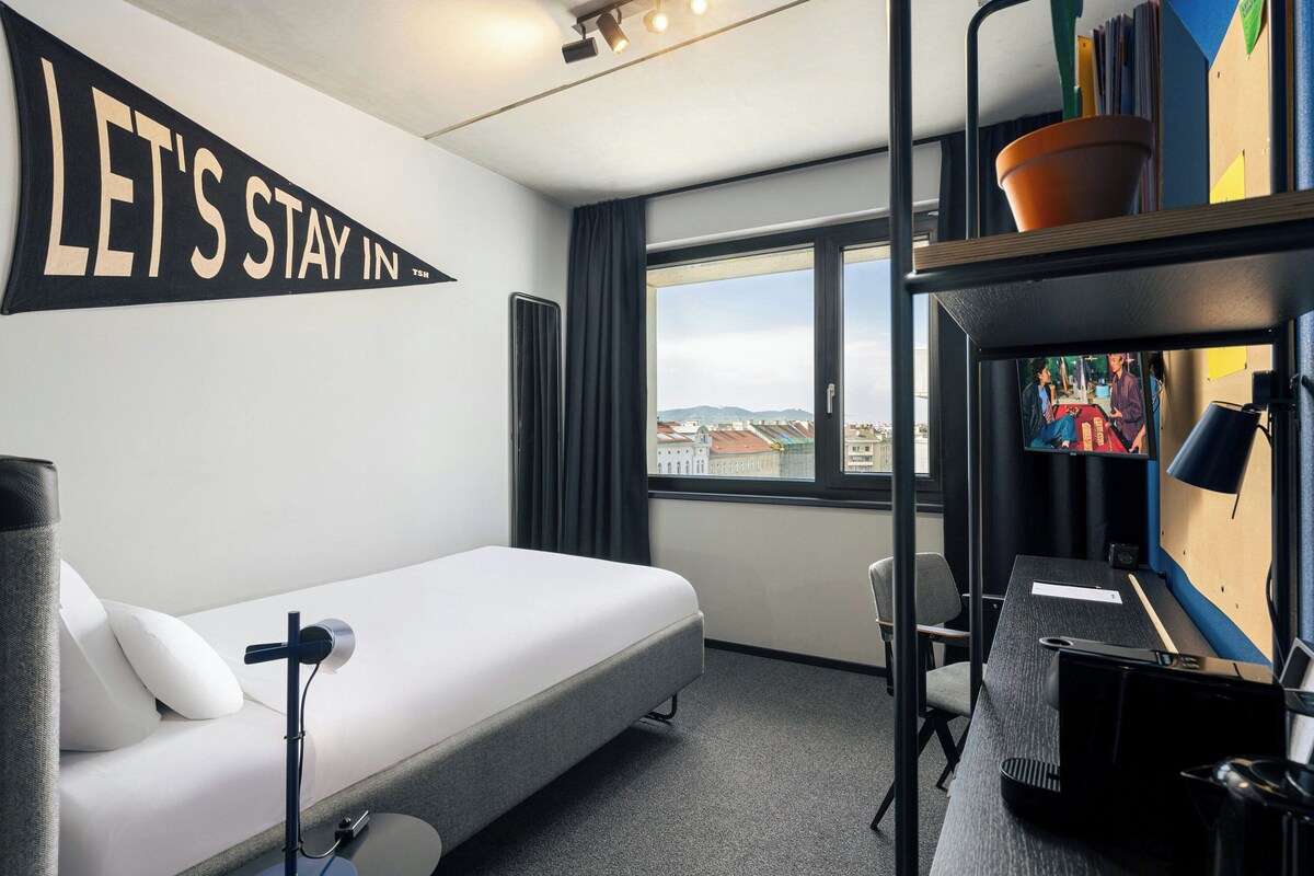 Habitación de hotel con cama, escritorio, silla, televisión y ventana con vista a la ciudad.