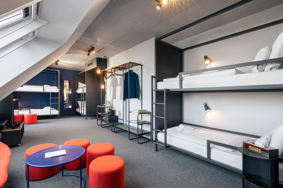 Une chambre d’hôtel moderne avec des lits superposés, un bureau et des poufs orange.