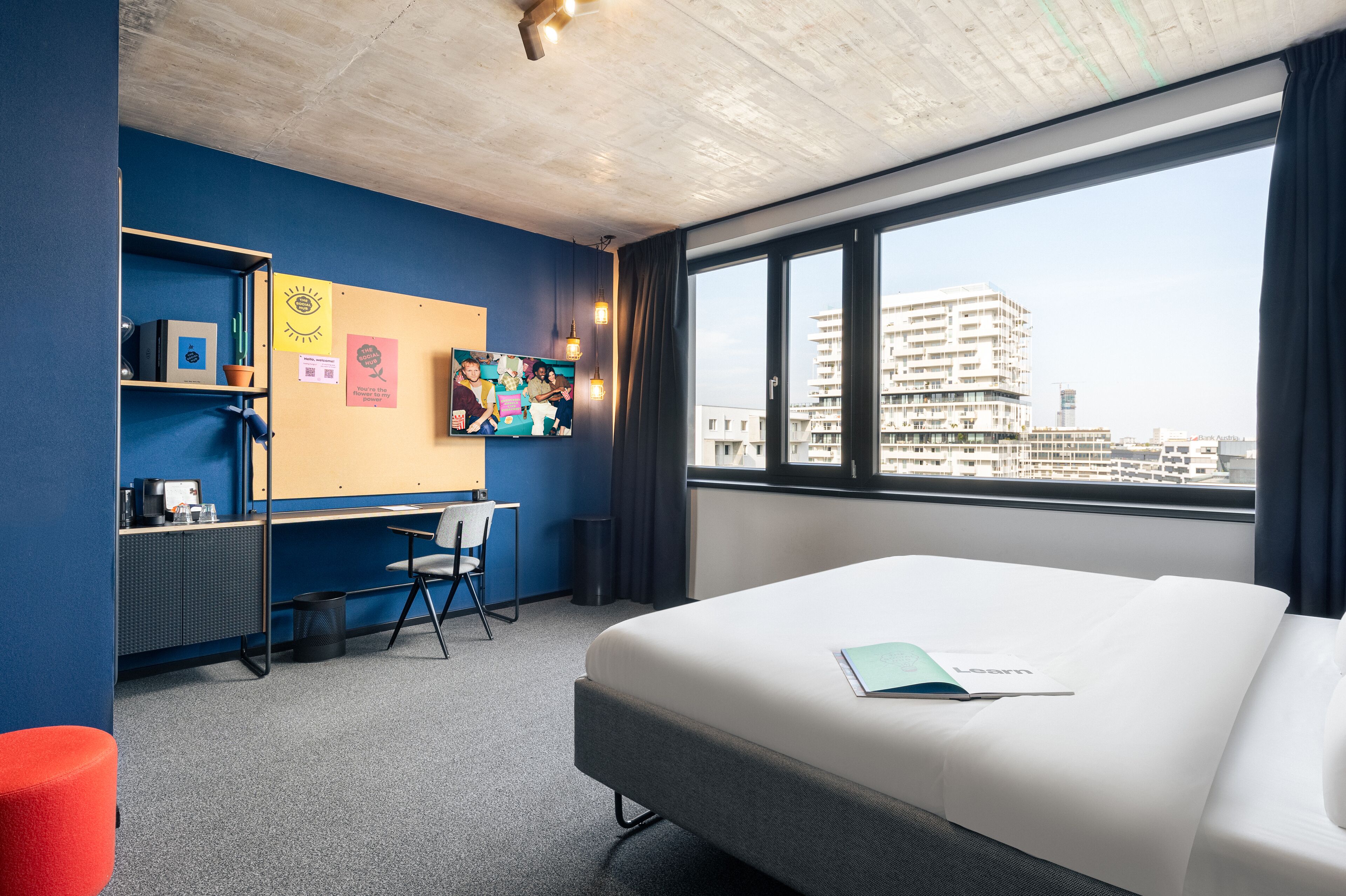 A hotel room with a bed, a desk, a chair, a bookshelf, and a large window with a city view.