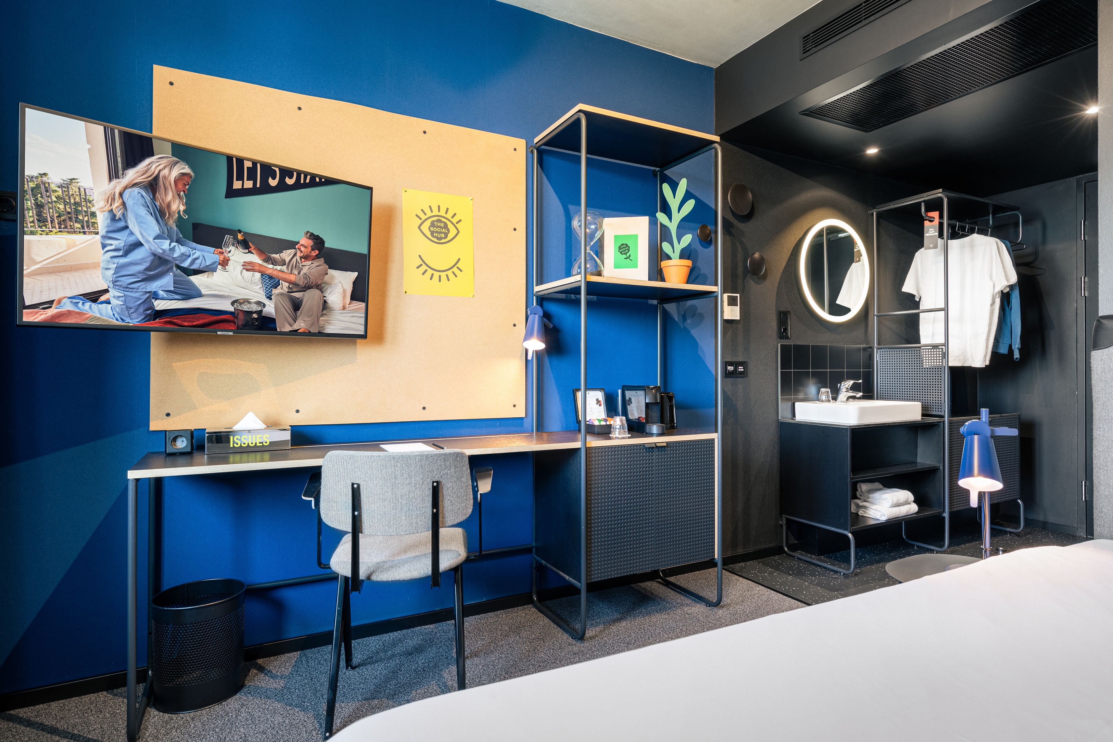 Ein modernes Hotelzimmer mit Schreibtisch, Sessel, Bett, Fernseher, Ablagefläche mit Pflanze, Spiegel und Waschbecken.