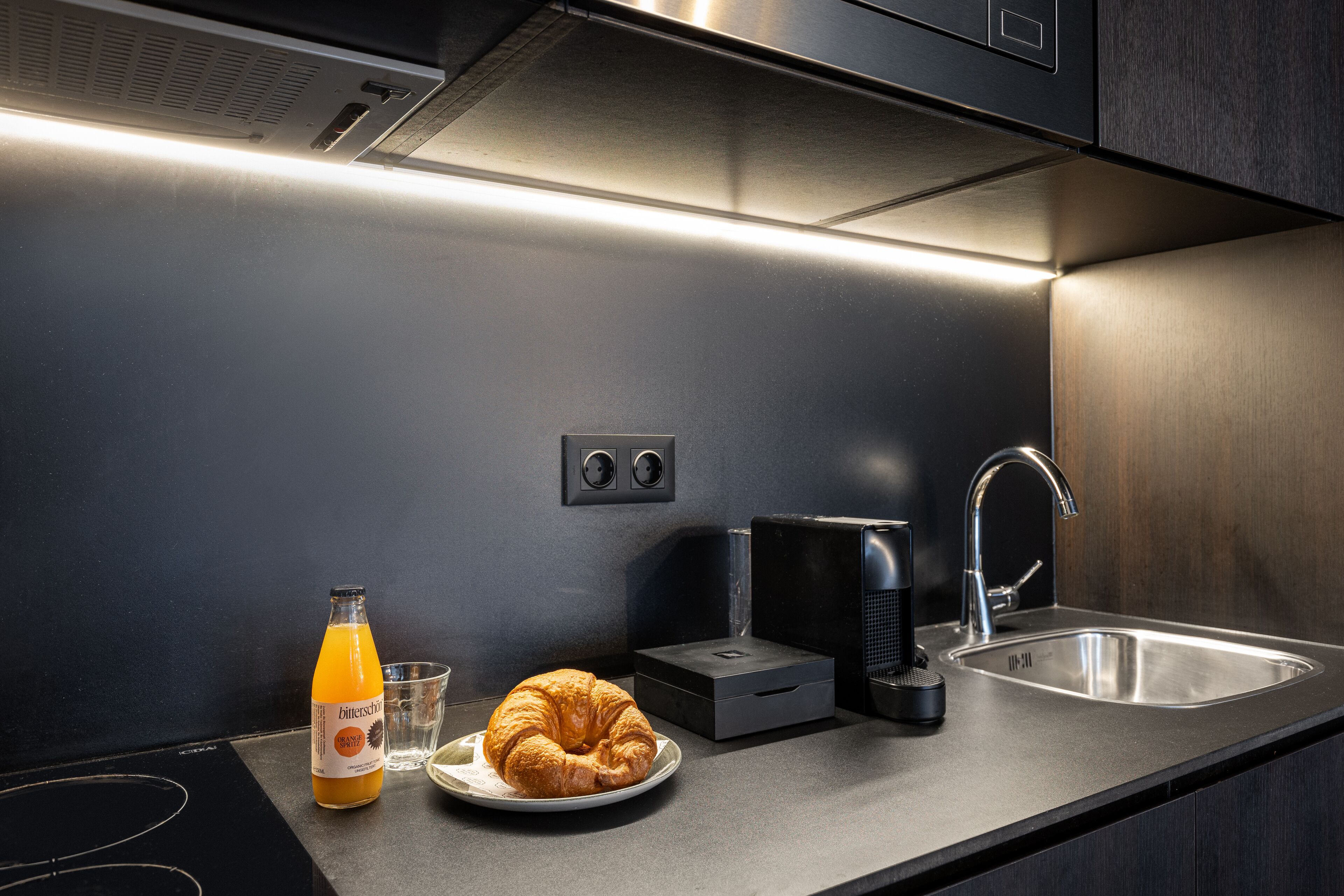 Uma cozinha moderna com uma pia de aço inoxidável, uma cafeteira, uma garrafa de suco de laranja e um croissant em um prato.