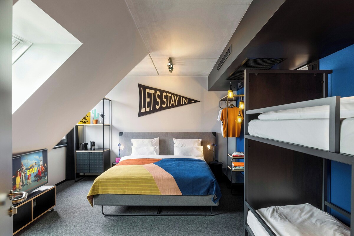 Une chambre d’hôtel moderne équipée d’un lit superposé, d’un téléviseur à écran plat, d’un bureau avec une chaise et d’une pancarte sur le mur indiquant « Restons à l’hôtel ».