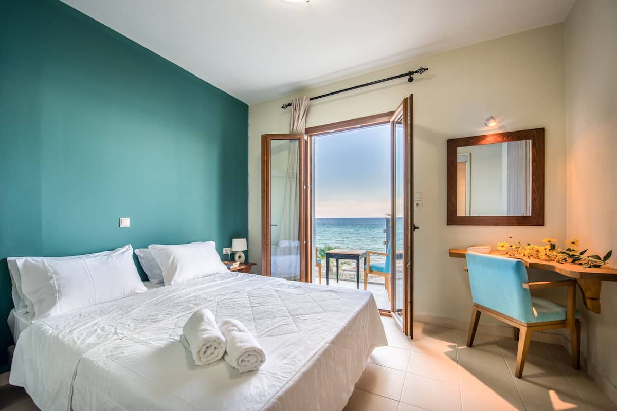 A bedroom with a bed, a desk, a chair, and a view of the sea.