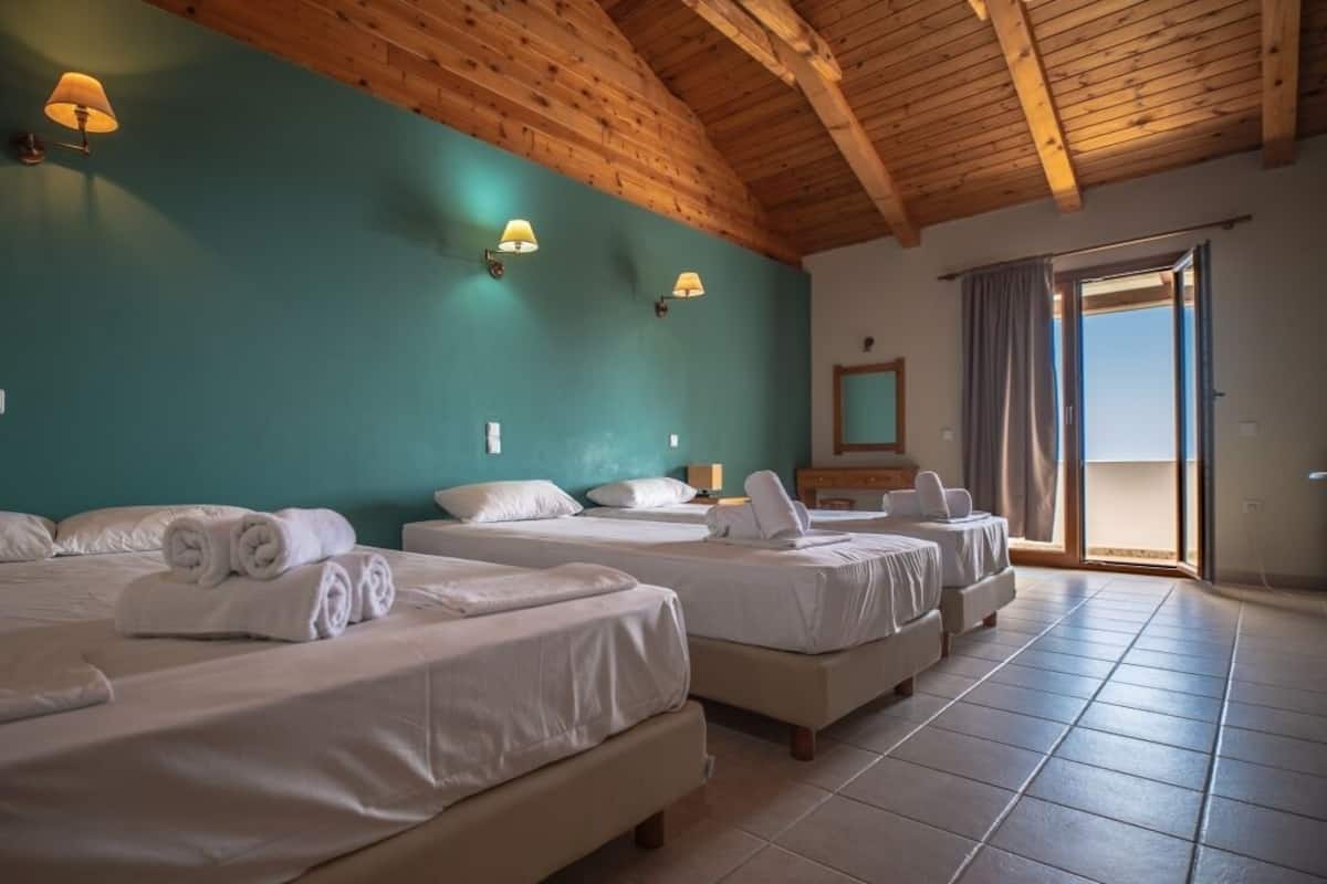 A hotel room with three beds, wooden ceiling beams, teal walls, and a sliding glass door to the outside.
