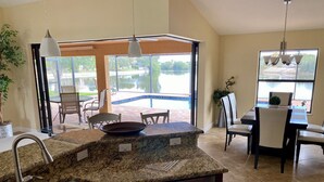 A kitchen with granite countertops, a dishwasher, and a sink. There is a dining area with chairs and a table, and a view of a swimming pool through large windows. - Southern Sun Beach Hotel - Cape Coral (Cape Coral)
