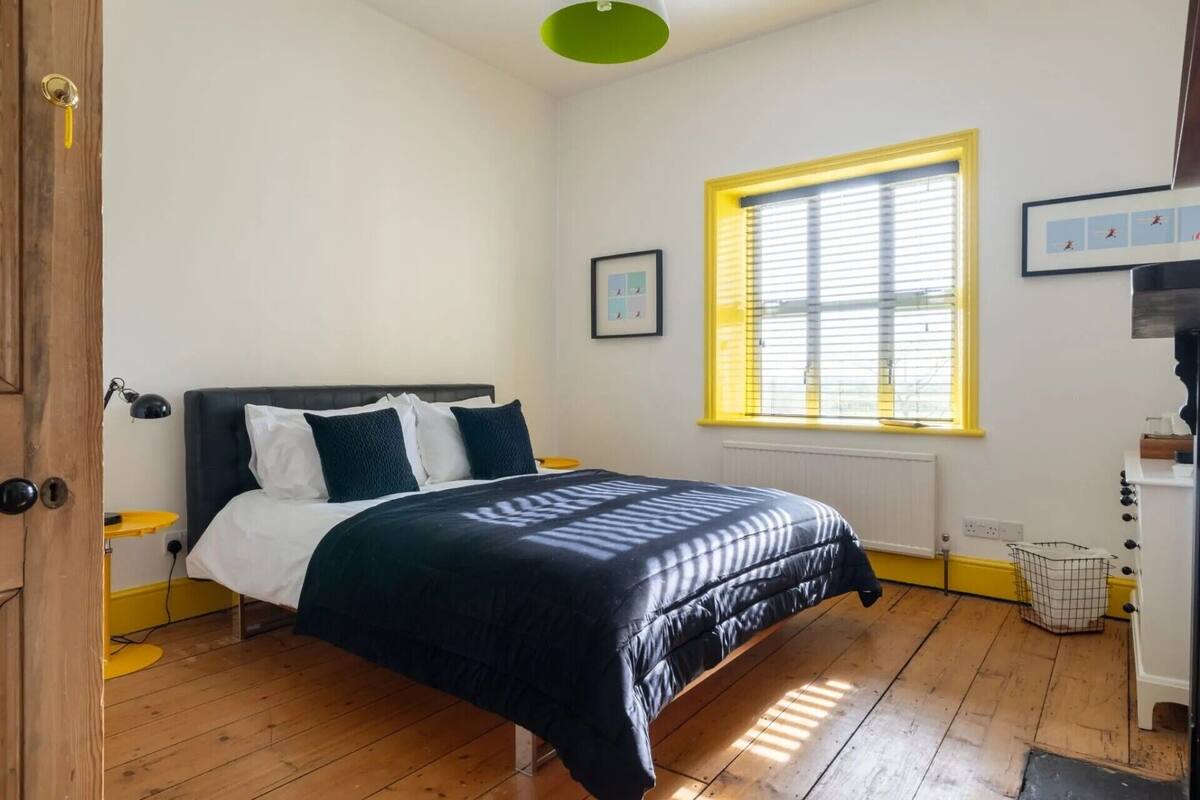 A bedroom with a bed, a window with blinds, a bedside table, and a wall-mounted picture.