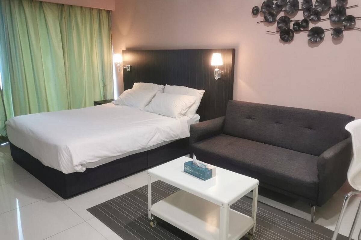 A hotel room with a bed, a grey sofa, a white bedside table, and a wall-mounted lamp.