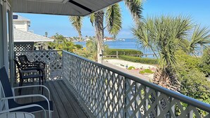 Une terrasse en bois avec des sièges d’extérieur, une balustrade blanche en treillis et une vue sur l’océan et les palmiers.