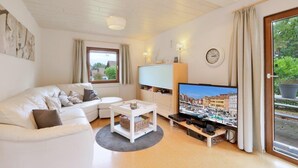 Un salon moderne avec un canapé blanc, un téléviseur et une grande fenêtre donnant sur des immeubles et de la verdure.