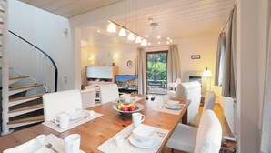 Un coin repas avec une table en bois dressée pour quatre personnes, des chaises blanches et un saladier rempli de fruits. On aperçoit un téléviseur au fond, et un escalier se trouve sur la gauche.