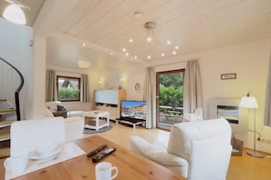 Una sala de estar moderna con techo de madera, un televisor de pantalla plana, una zona de comedor con mesa y sillas, y una chimenea.