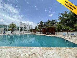 A swimming pool with lounge chairs and a wooden structure, surrounded by palm trees and a building with large windows.