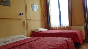 A room with two beds, each with a red bedspread and white pillows. There is a window with sheer curtains and a radiator on the wall. - Hôtel du Palais (Rouen)