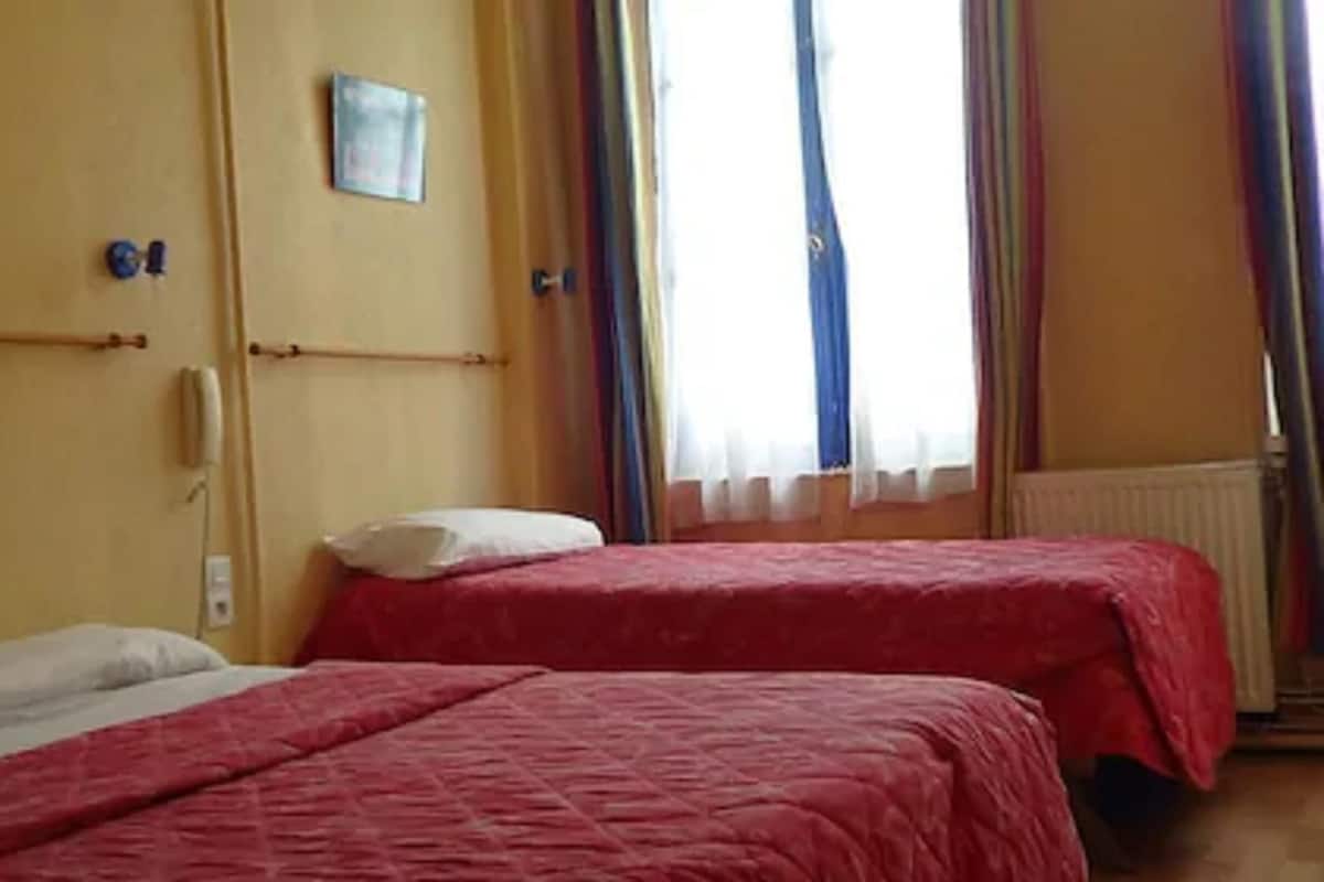 Una habitación con dos camas, cada una con una colcha roja y almohadas blancas. Se ve una ventana con cortinas translúcidas y un radiador en la pared.