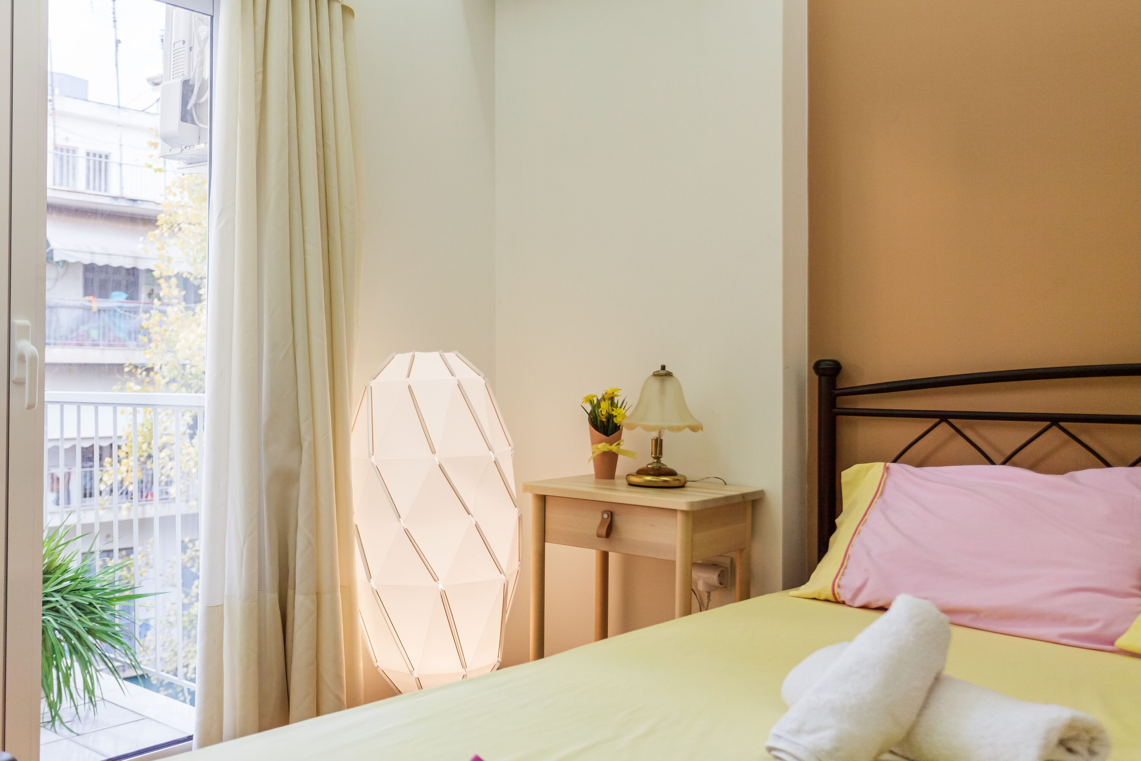 Une chambre d’hôtel avec un lit, une table de chevet avec une lampe et des fleurs, une fenêtre avec des rideaux et une vue sur les bâtiments à l’extérieur.