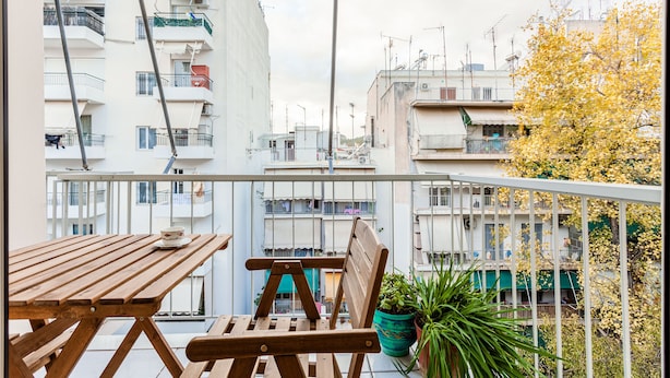 A balcony with a wooden table and chair, potted plants, and a view of surrounding buildings. - Zografou Hidden Gem - Athens Charming Home (Zografos)
