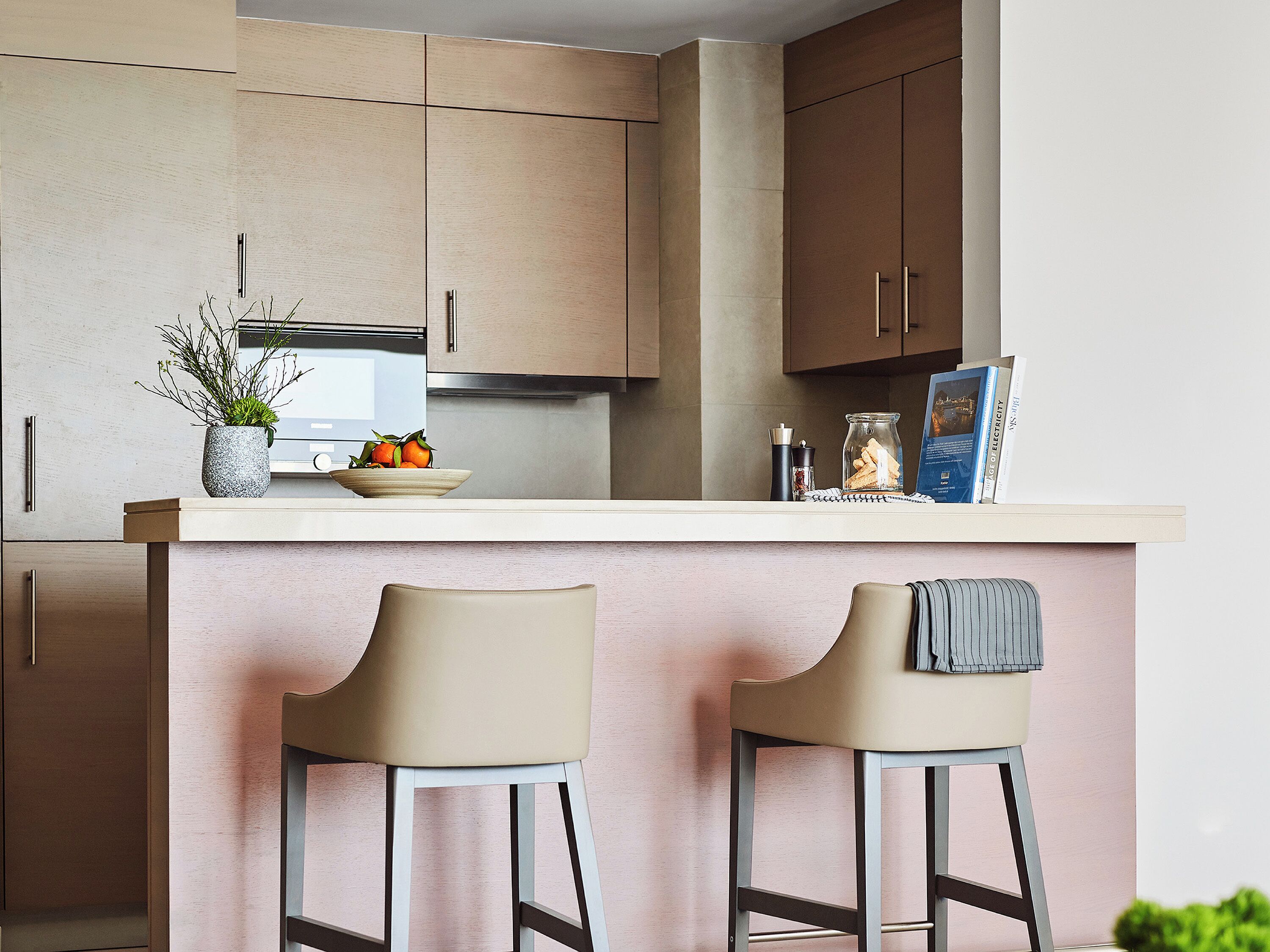 A modern kitchen with two bar stools, a countertop with a bowl of fruit, a potted plant, and a book.
