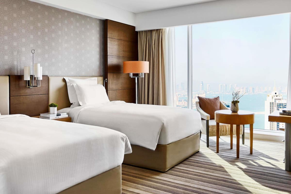 A hotel room with two beds, a nightstand, a lamp, a chair, and a view of the city.