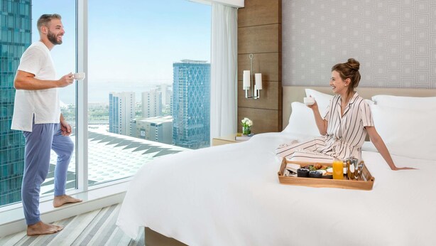 Une chambre d’hôtel avec une grande fenêtre, un lit sur lequel se trouve un plateau avec des articles pour le petit-déjeuner, et une table de chevet ornée d’un vase de fleurs.
