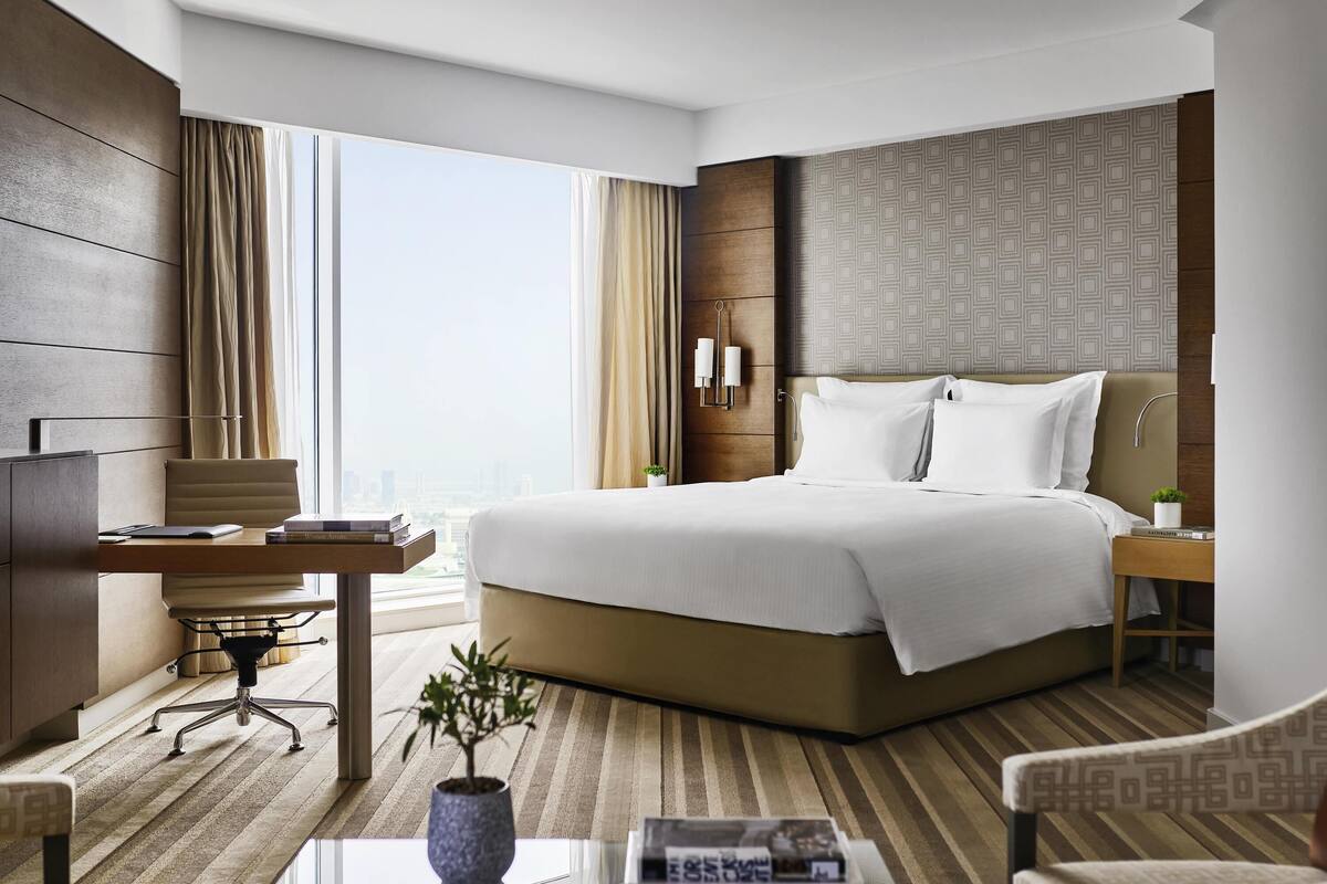 A hotel room with a large bed, a desk, a chair, a small table with a plant, and a view of the city.