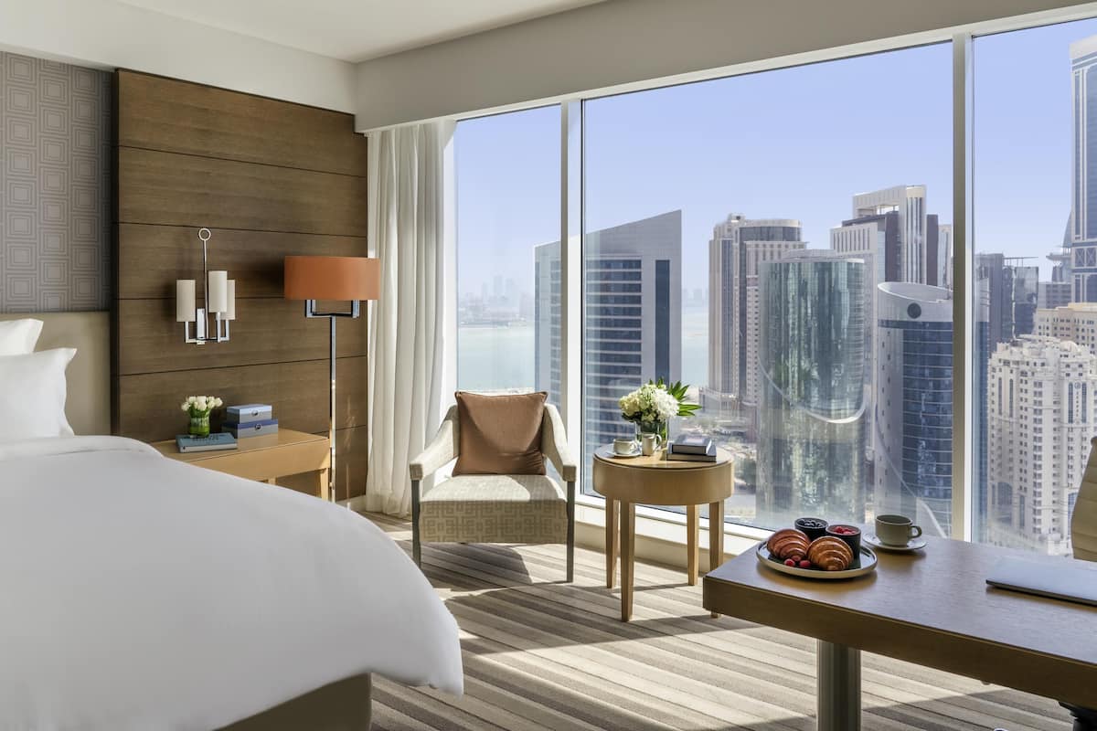 A hotel room with a large window, a bed, a chair, a small table with pastries, and a lamp.