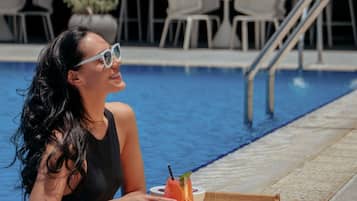Una persona sentada junto a una piscina, con una bandeja que contiene una bebida y un bol, rodeada de sillas y una escalera.