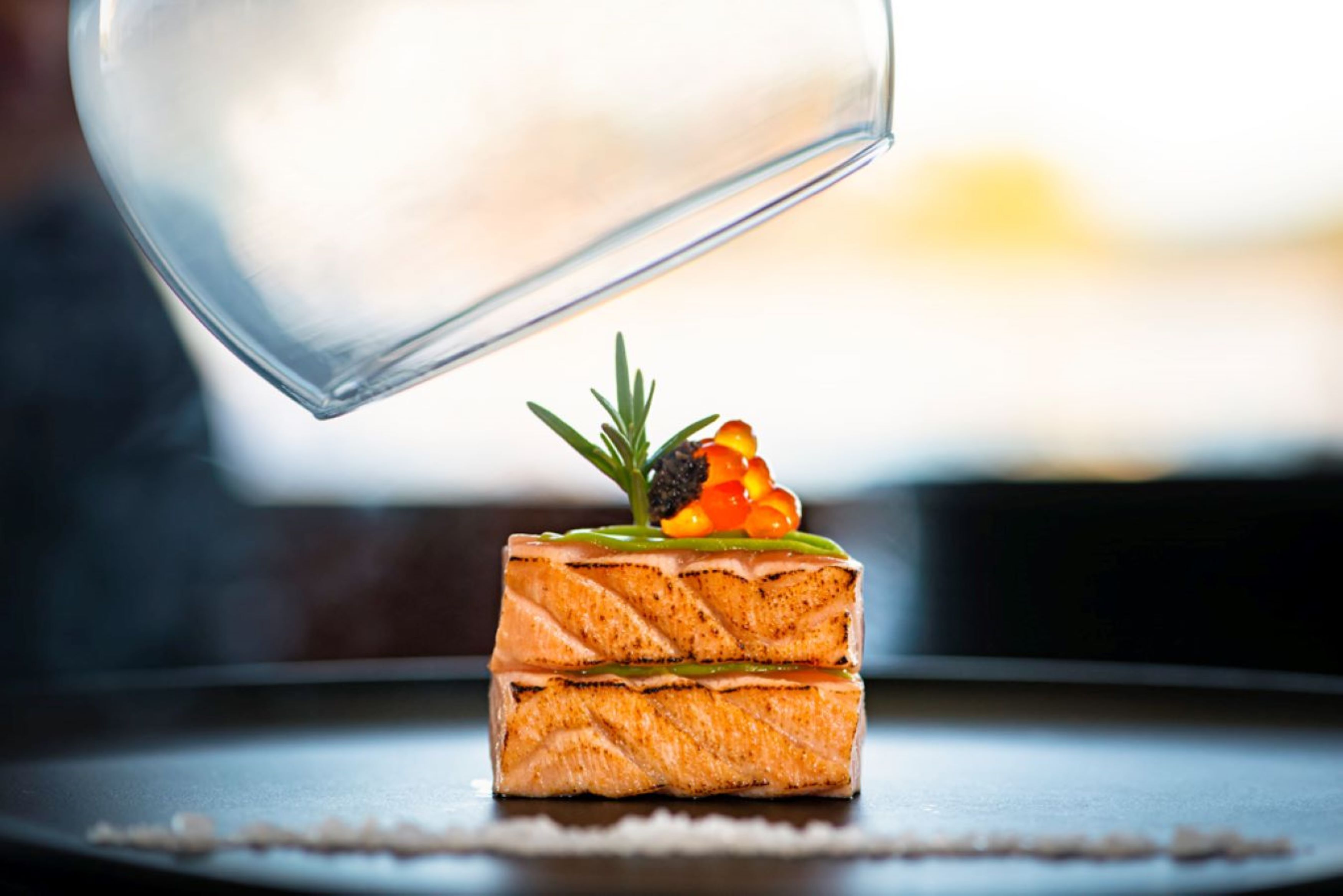 A glass of water being poured onto a salmon tartare dish with a garnish.
