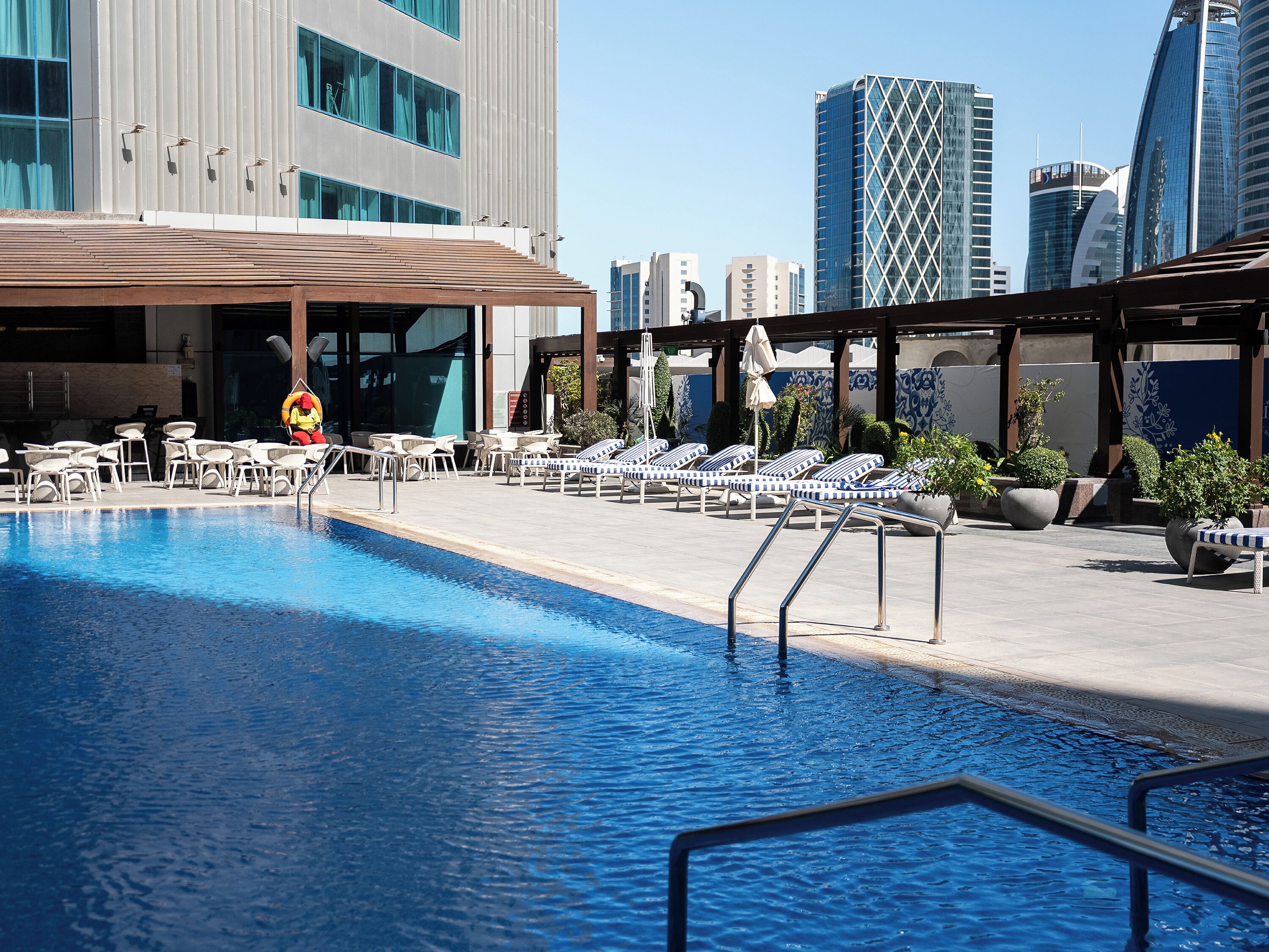 A swimming pool with lounge chairs and umbrellas, a lifeguard, and modern buildings in the background.