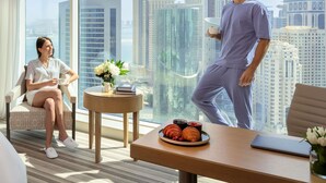 Une chambre d’hôtel moderne avec une grande fenêtre, une table ronde sur laquelle se trouve un ordinateur portable, une assiette de pâtisseries et un vase rempli de fleurs.