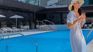 Un espace piscine doté de chaises longues et de parasols, une piscine à l’eau d’un bleu limpide et, en arrière-plan, des bâtiments modernes.