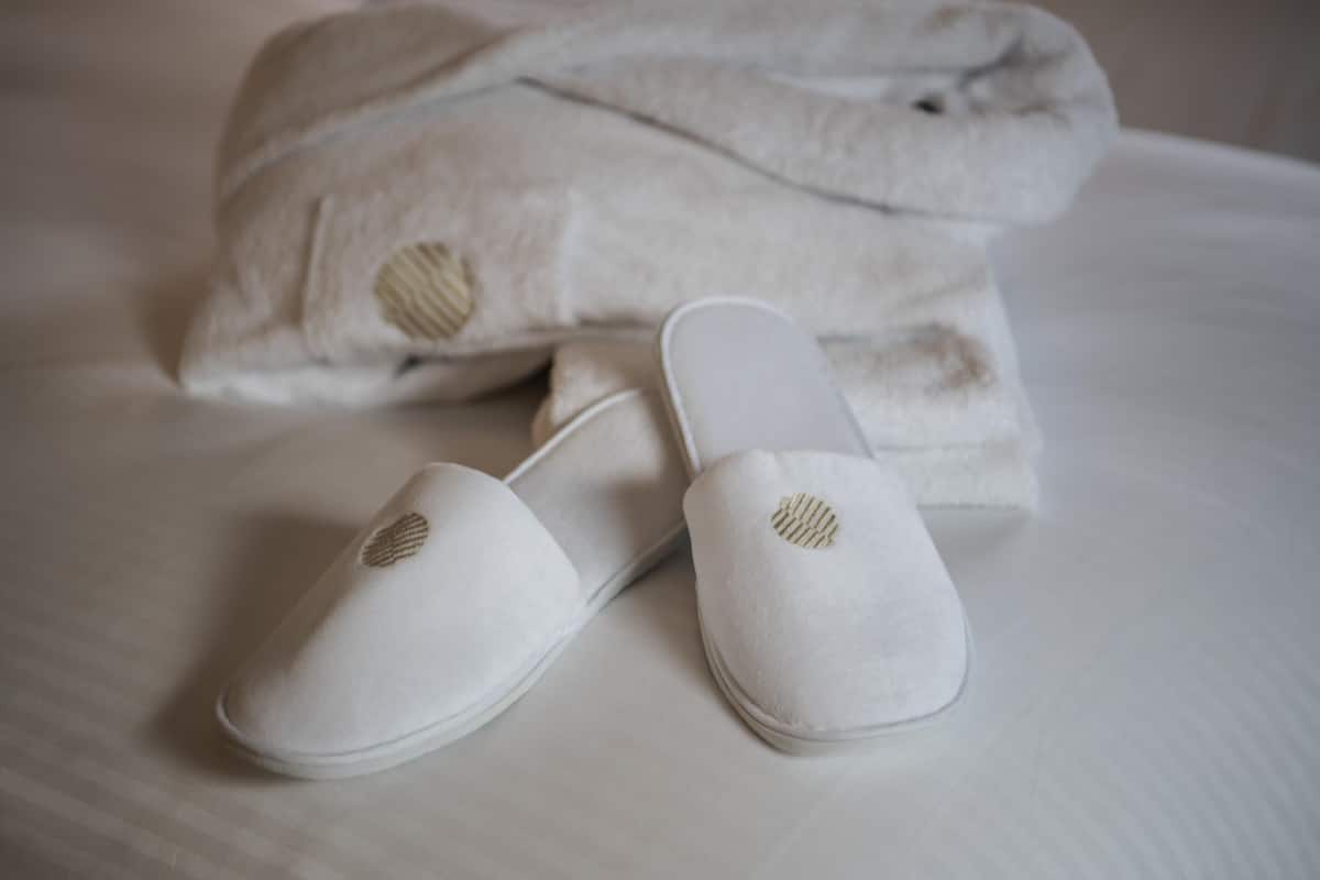 Una cama bien hecha con un almohadón blanco y un par de zapatillas blancas con un emblema dorado.