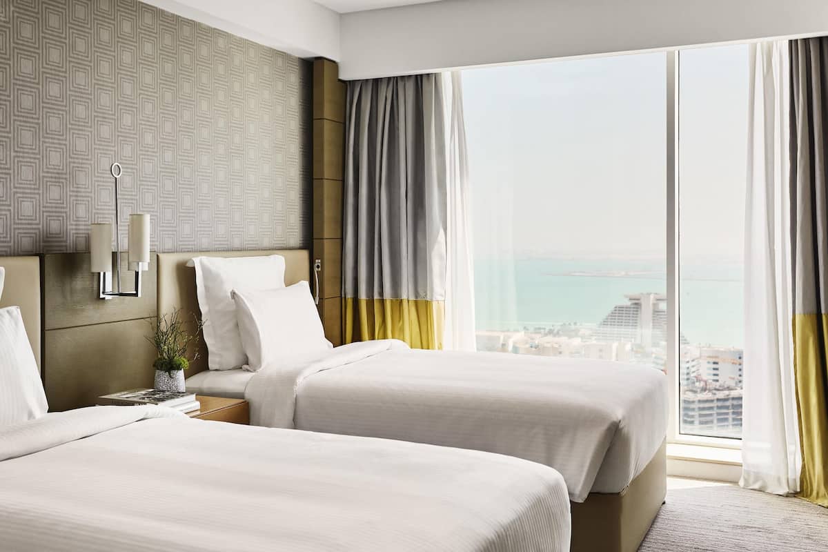 Habitación de hotel con dos camas, un ventanal con cortinas, un papel tapiz estampado y vistas a la ciudad.