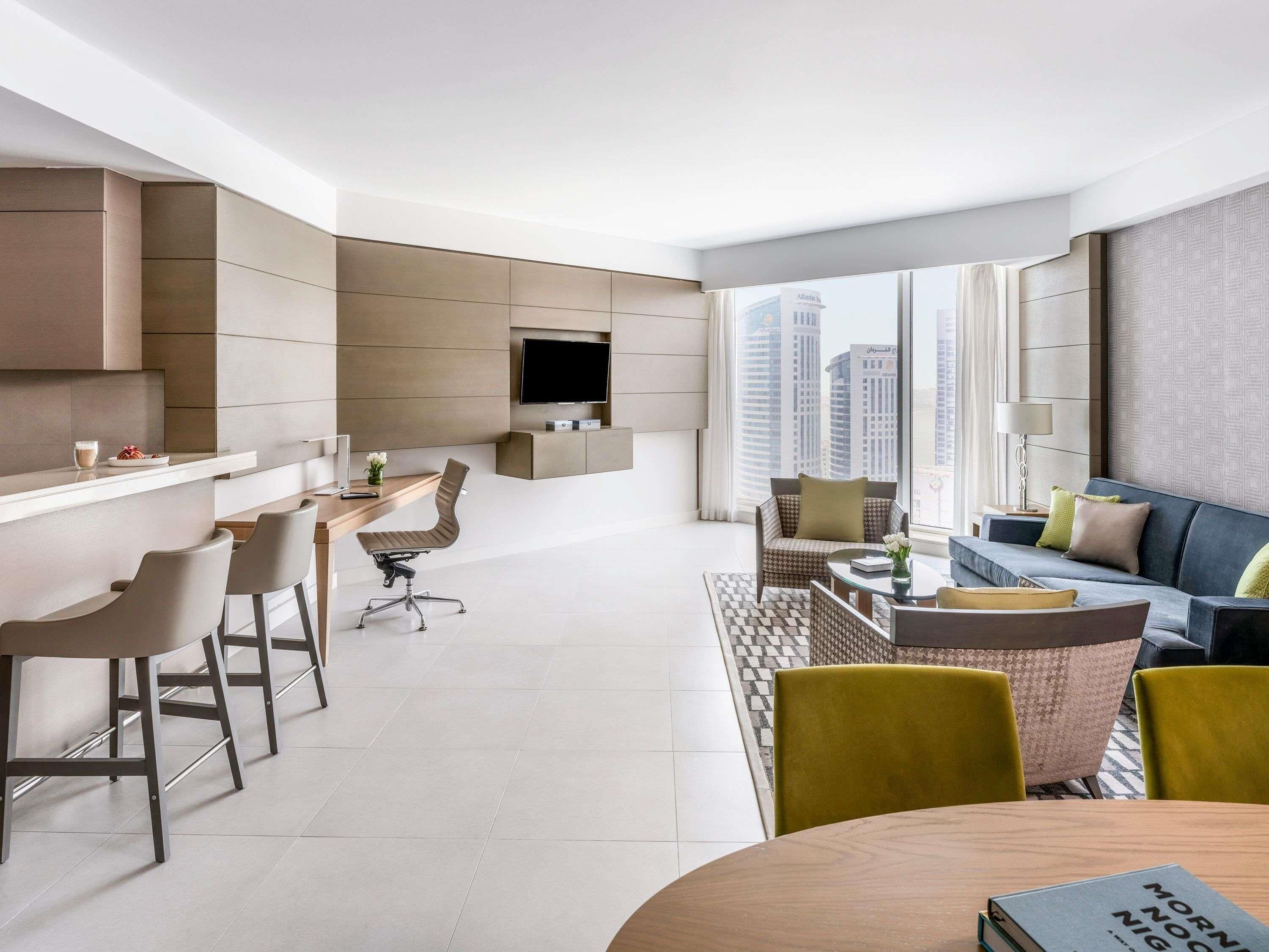 Una sala de estar moderna con un sofá, un comedor con sillas, un televisor montado en la pared y vistas urbanas a trav és de grandes ventanales.