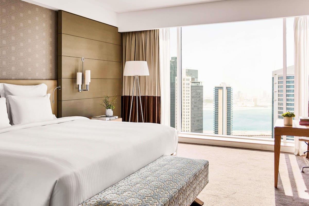 A hotel room with a large bed, bedside lamps, a bedside table with a plant, a floor lamp, and a view of the cityscape.