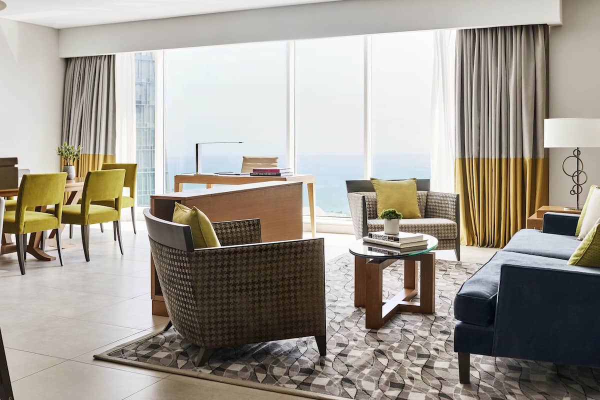 Una habitación de hotel moderna con un ventanal grande, un sofá, una silla, una mesita y un escritorio.