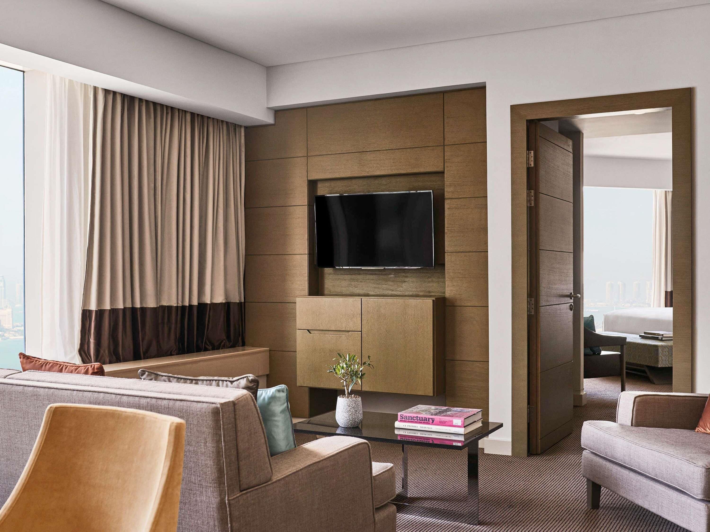 A modern hotel room with a large window, a flat-screen TV, a sofa, a coffee table, and a door leading to another room.