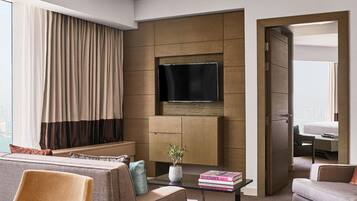 A modern hotel room with a large window, a flat-screen TV, a sofa, a coffee table, and a door leading to another room.