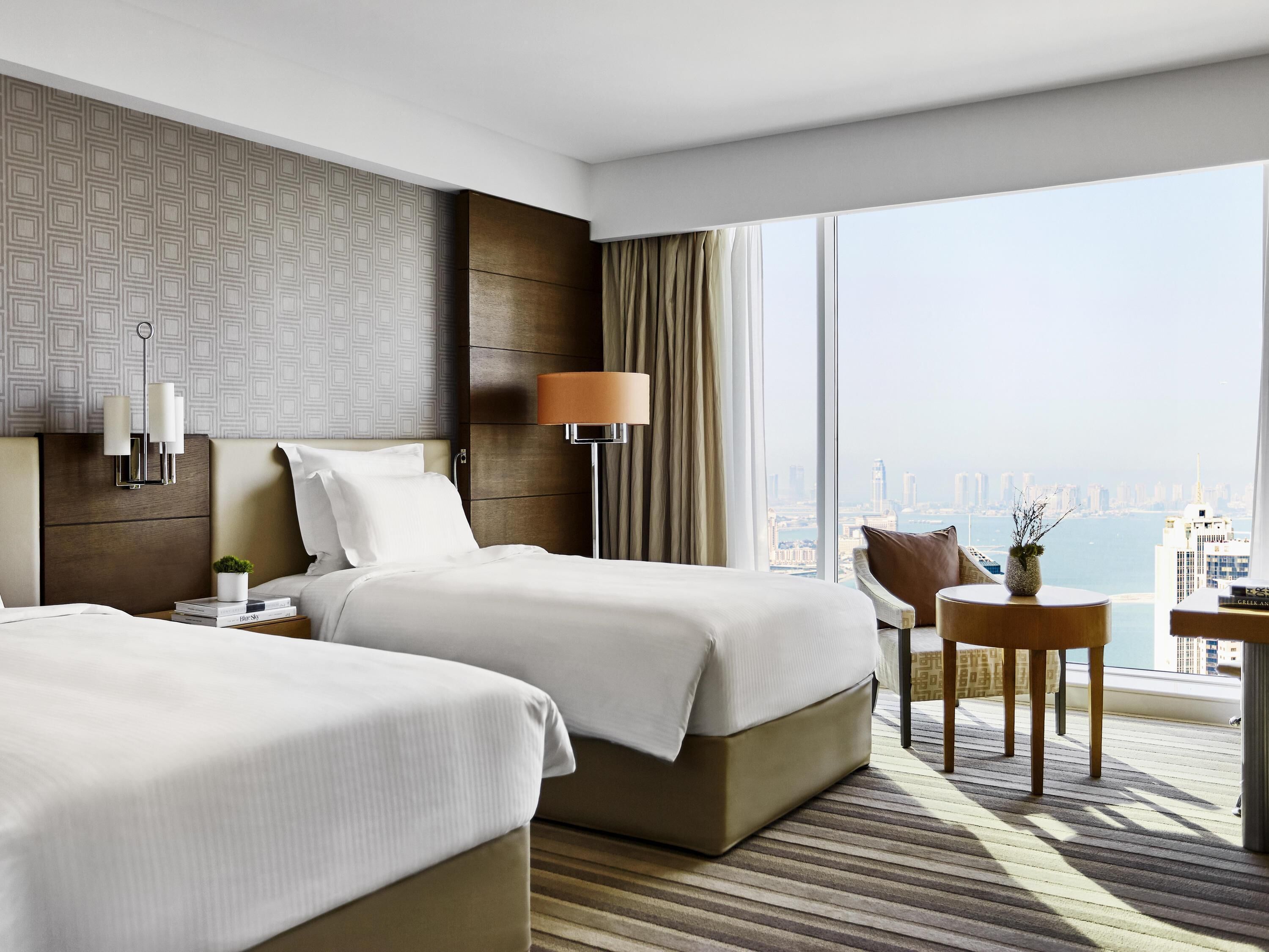 Une chambre d’hôtel avec deux lits, une table de chevet, une lampe, une chaise, une petite table et une vue sur la ville.