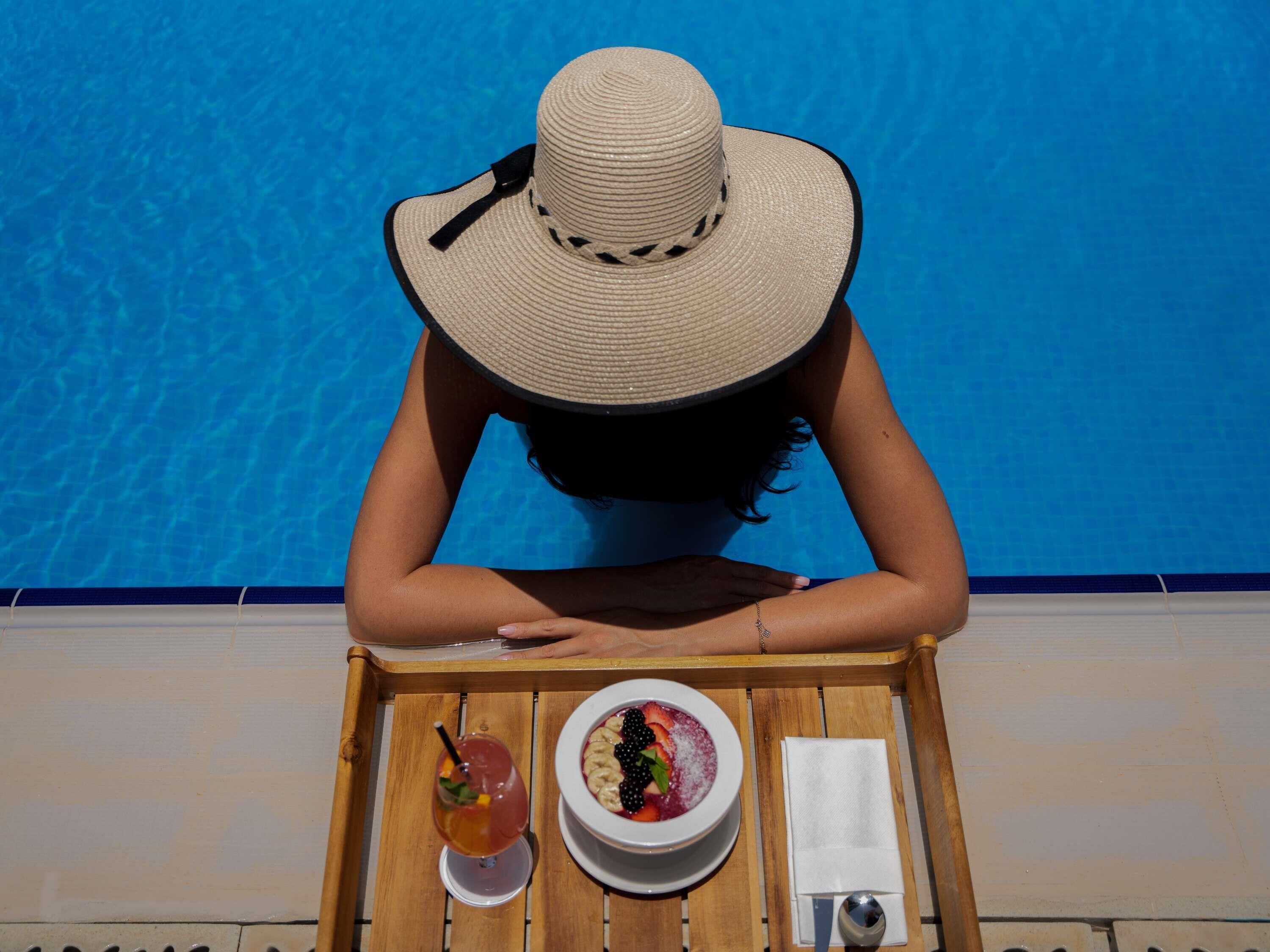 A person seated at a wooden table by a pool, with a drink and a bowl of fruit.
