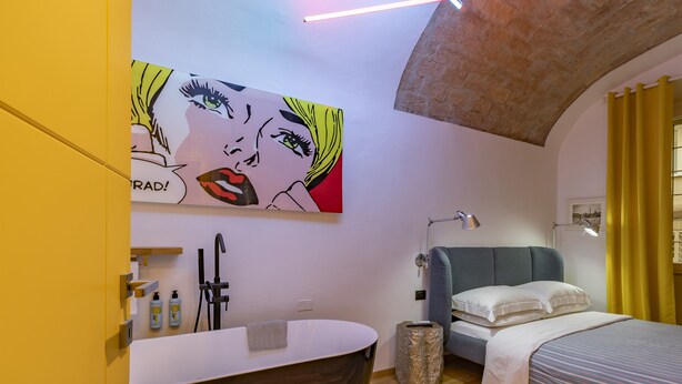 Un baño moderno con una bañera independiente, ducha y lavamanos. Hay una cama con cabecera azul y una lámpara de noche. Un cuadro grande de arte pop adorna la pared sobre la cama.