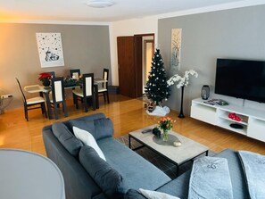 A modern living room with a grey sofa, a coffee table, a TV, a Christmas tree, and a dining area with chairs and a table.