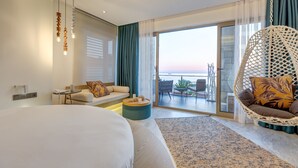 A modern hotel room with a large bed, a sofa, a hanging chair, and a balcony with a view of the sunset.