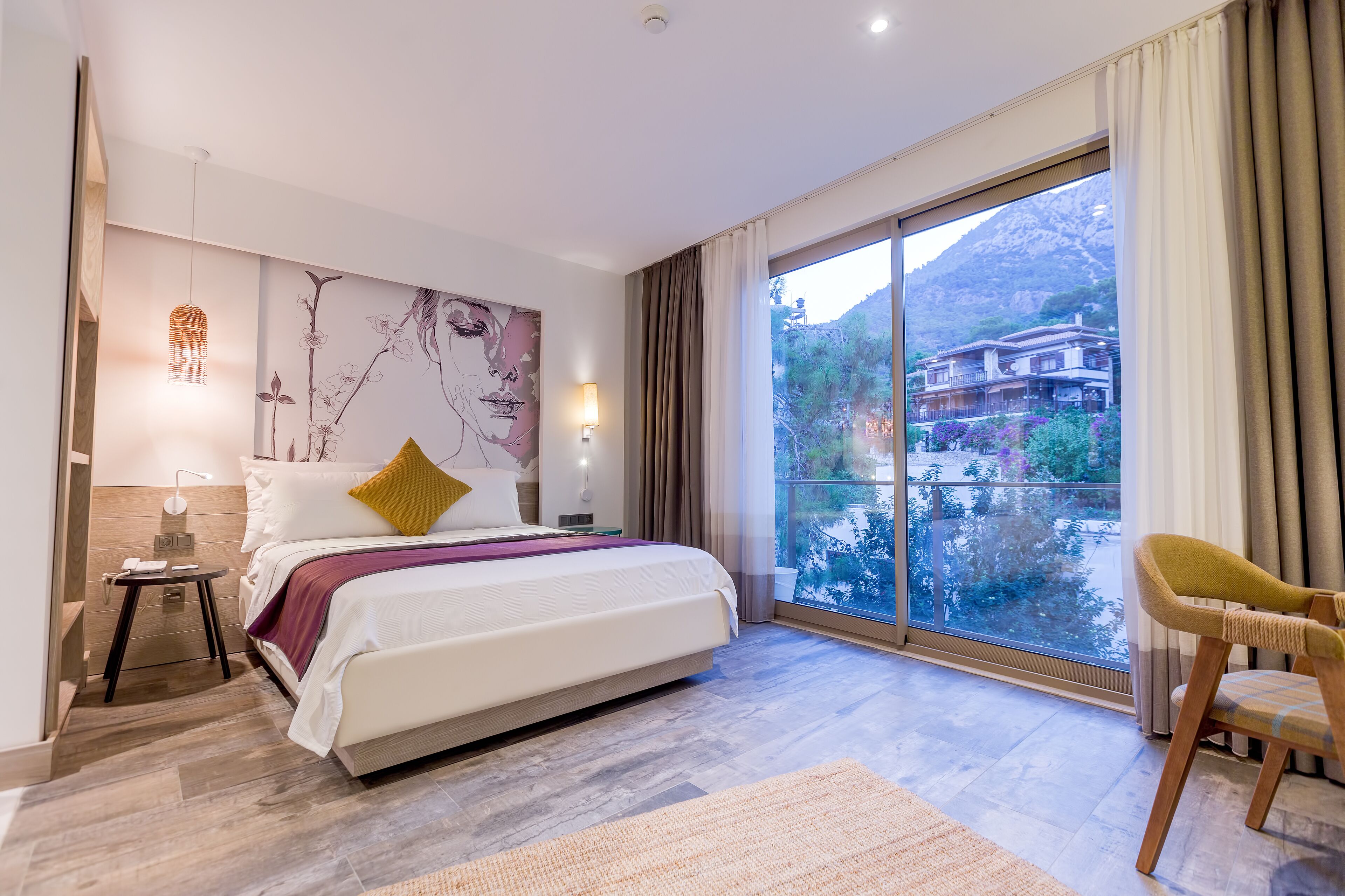 A modern hotel room with a large bed, a side table, a lamp, a chair, and a large window with a mountain view.