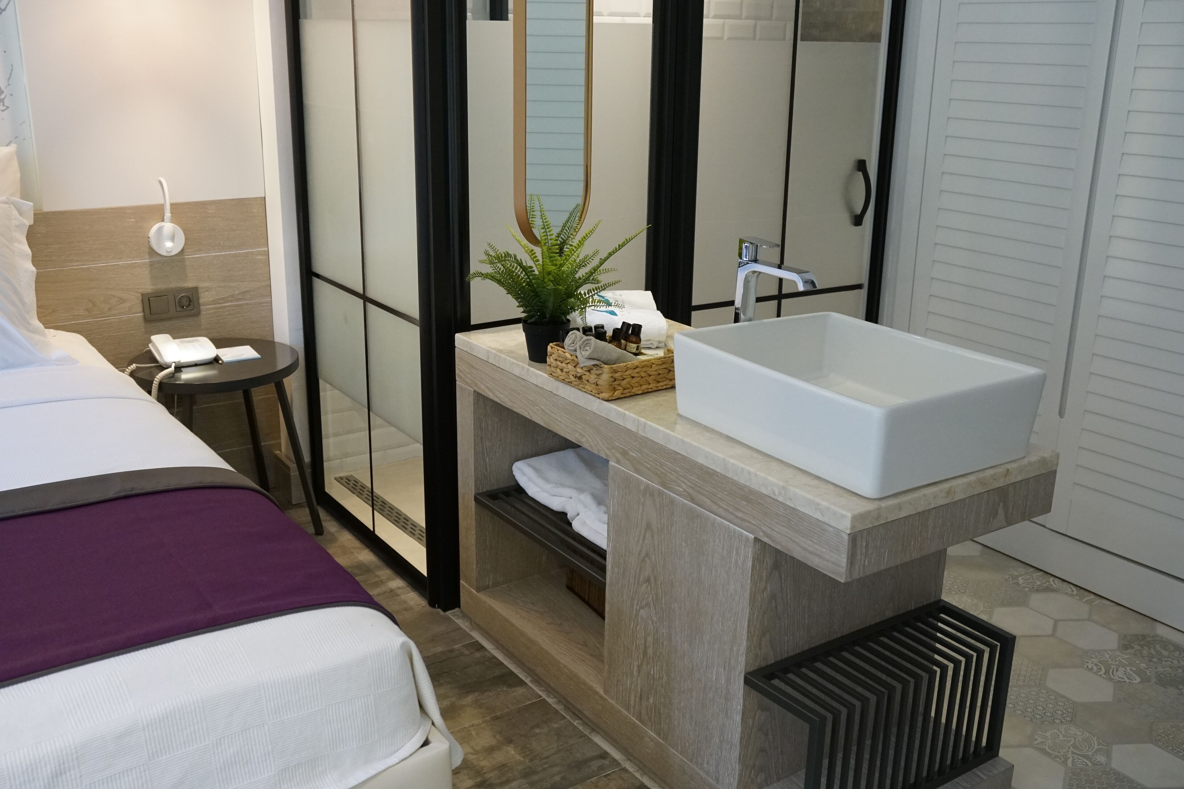 A modern hotel room with a large bed, a bedside table with a phone, a small table with a plant, a large mirror, and a built-in sink.