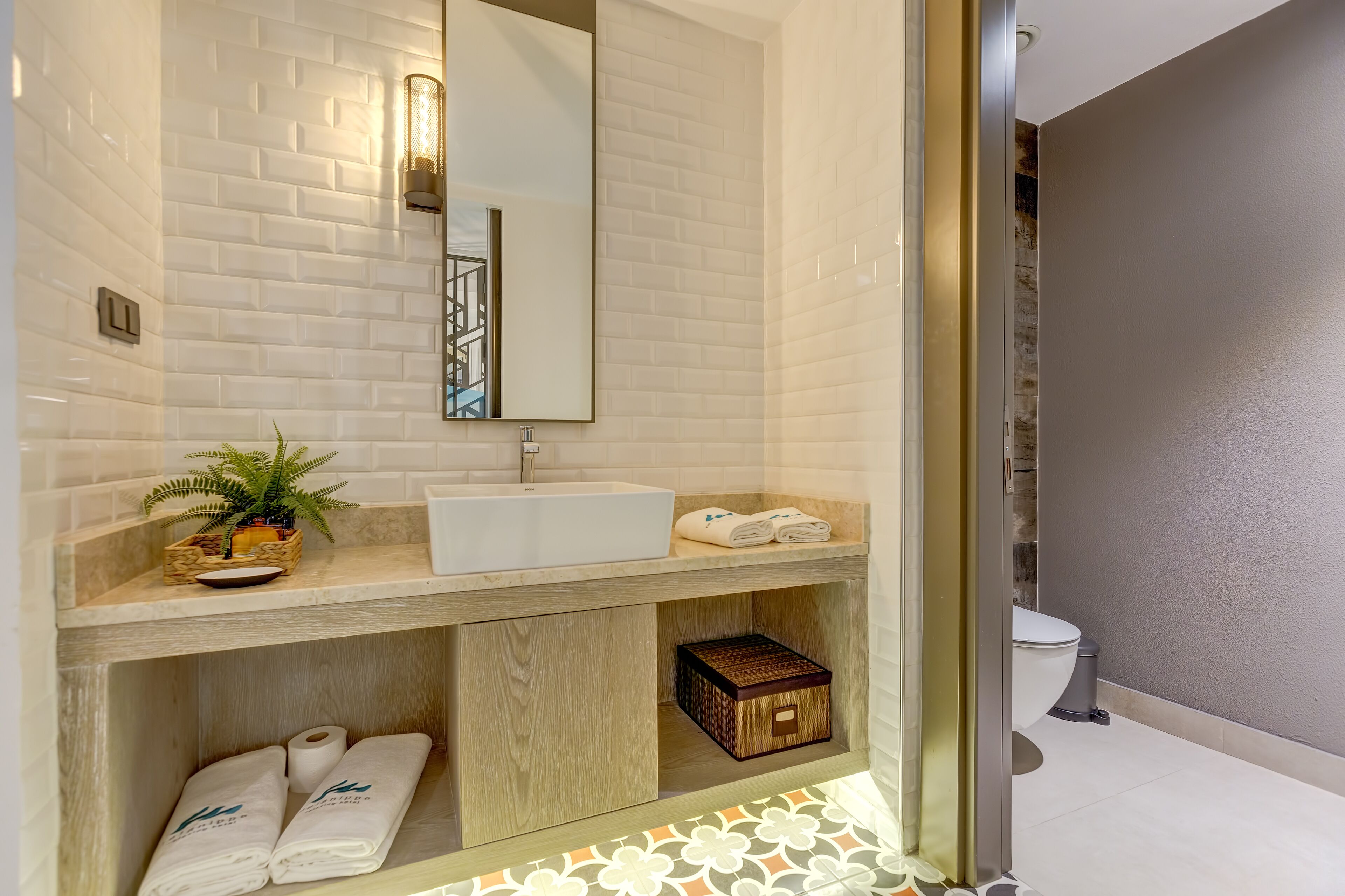A modern bathroom with a white sink, a mirror, and a potted plant. There are towels and a small wooden box on the counter.