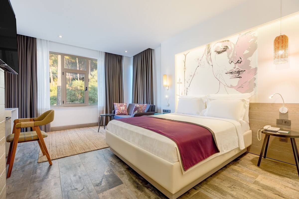 Habitación de hotel moderna con una cama grande, un escritorio y una silla. Hay una ventana con cortinas, una lámpara de pared y un cuadro en la pared.