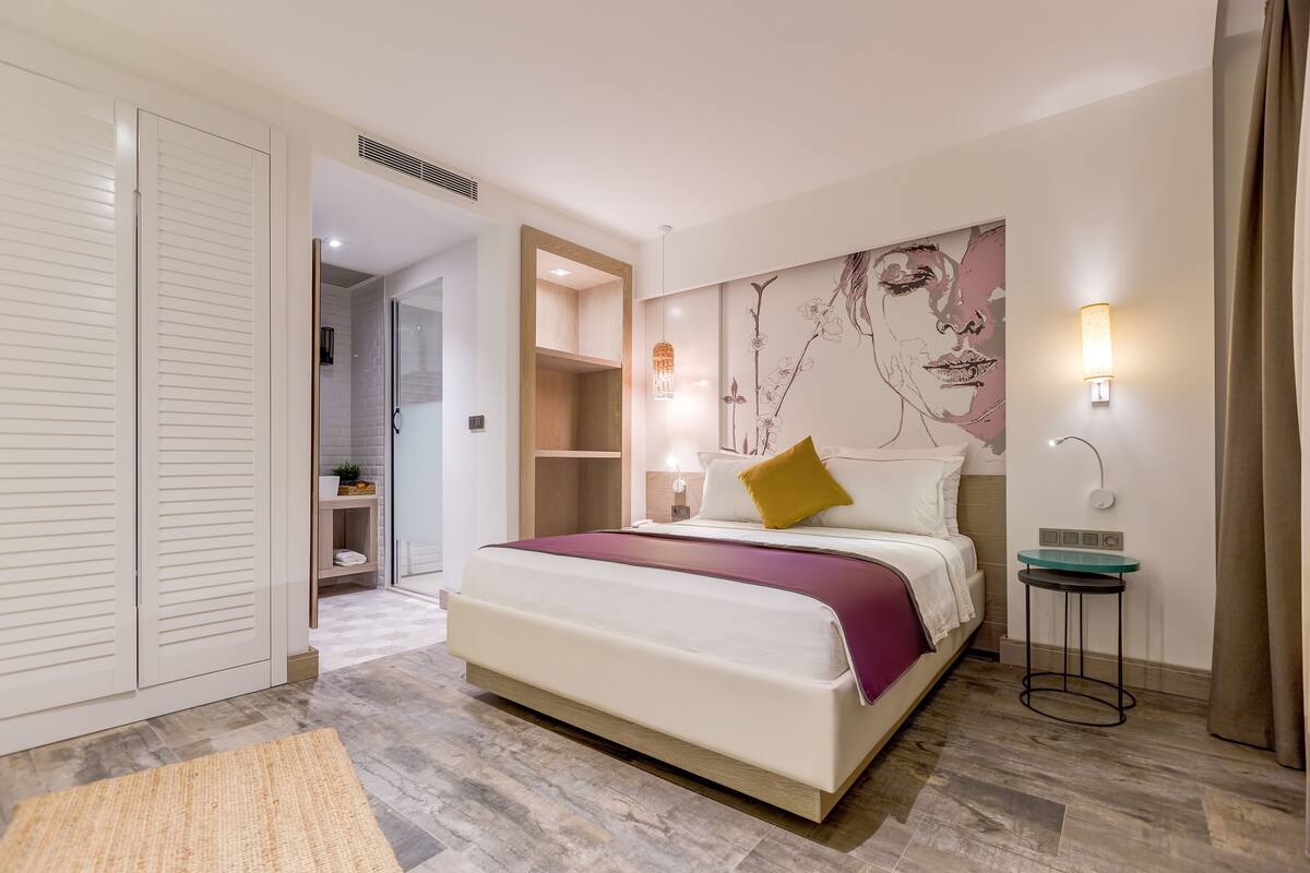 Una habitación de hotel moderna con una cama grande, un armario, una mesita y una lámpara de pared.