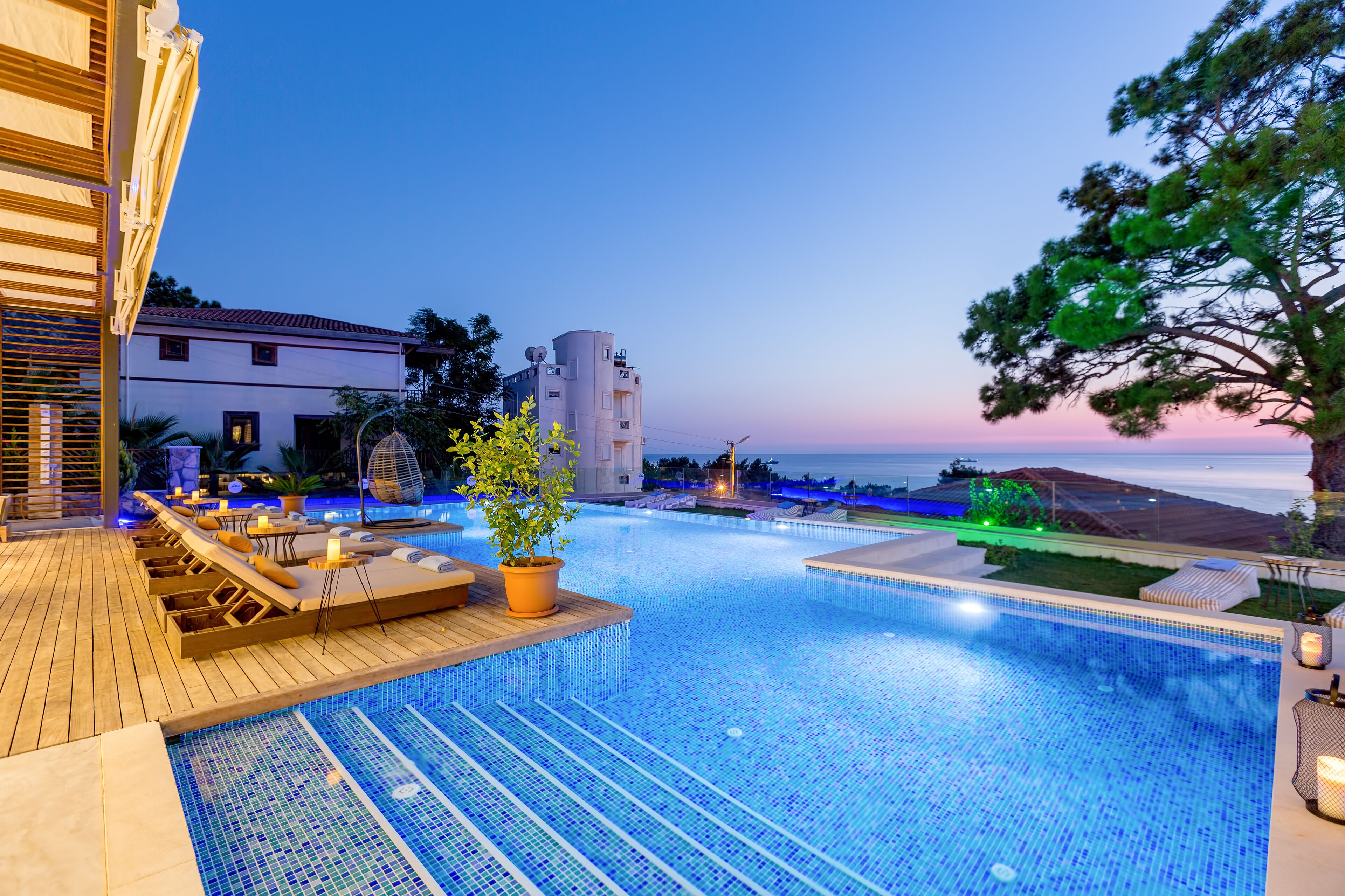 A poolside area with lounge chairs, a potted plant, and a view of the sea at dusk.